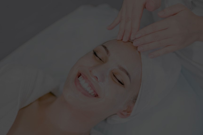 Person lying down with eyes closed, a towel wrapped around their head, receiving a facial treatment.